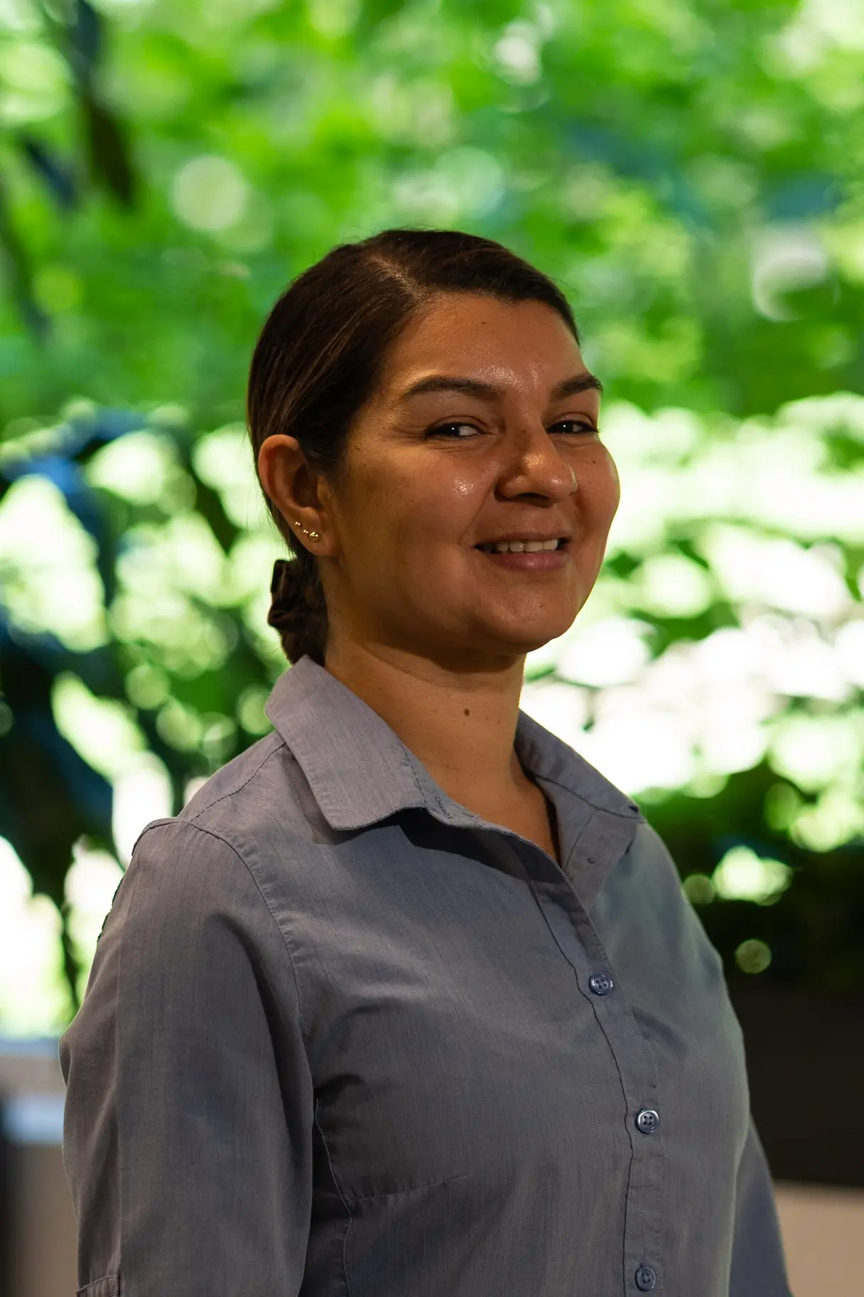 Woman with her hair in a tight bun, wearing a grey shirt and smiling against a natural background.