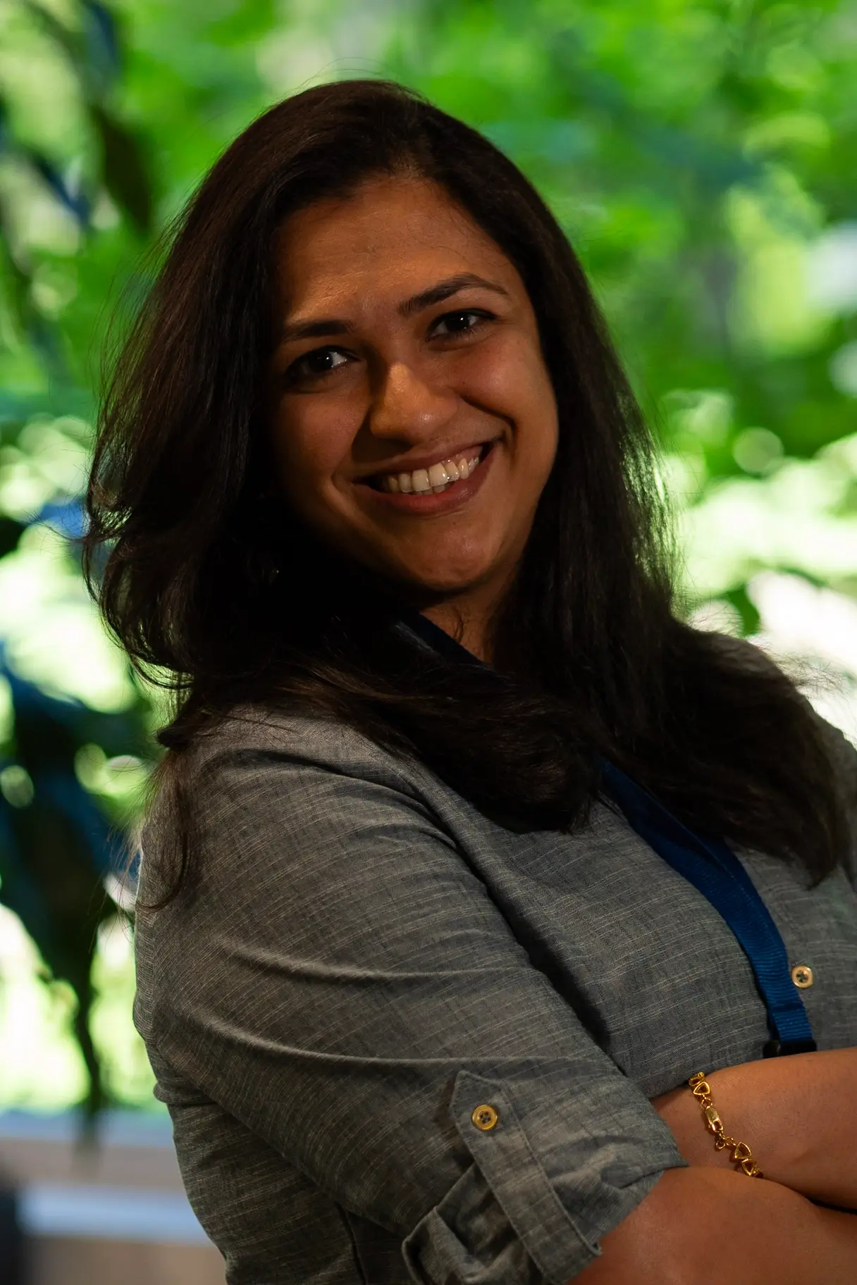 Woman with her hair down, wearing a grey dress and smiling against a natural background.