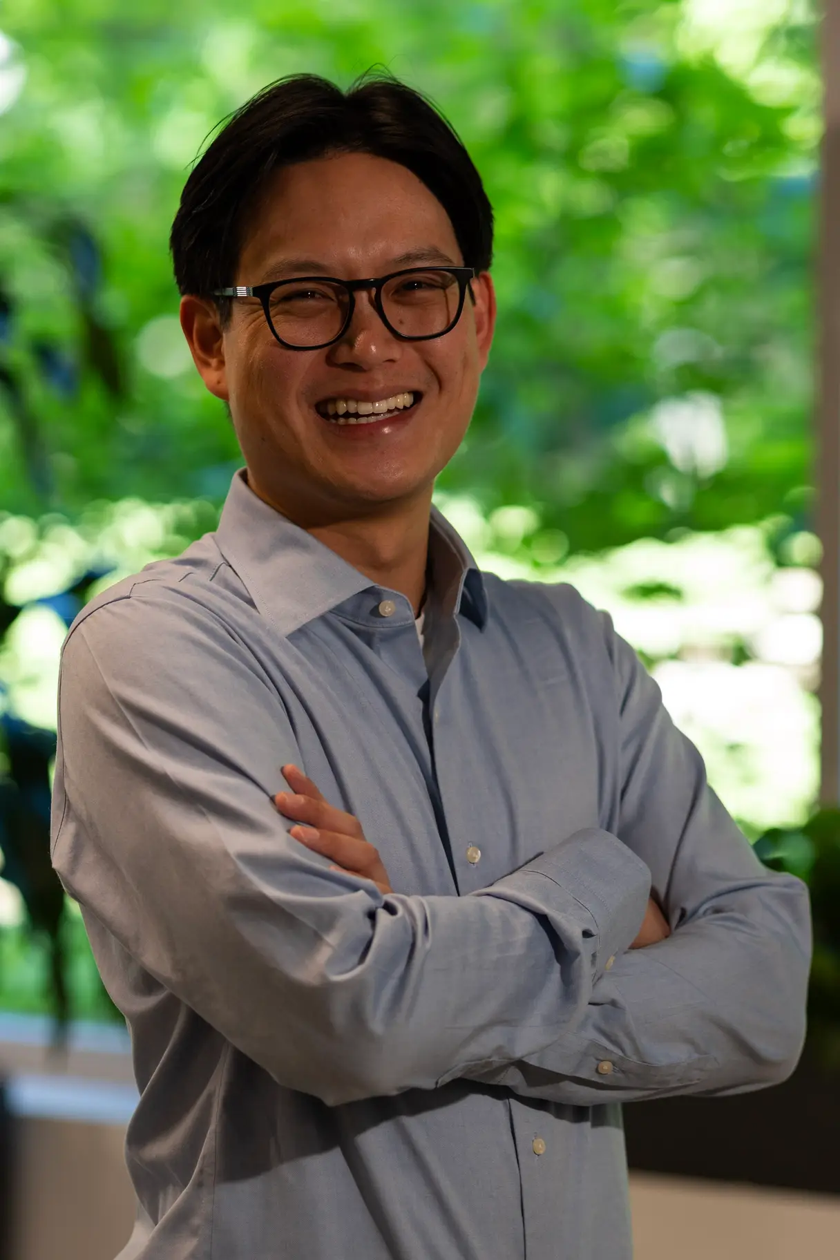Man in a light blue shirt wearing glasses, smiling against a natural background.