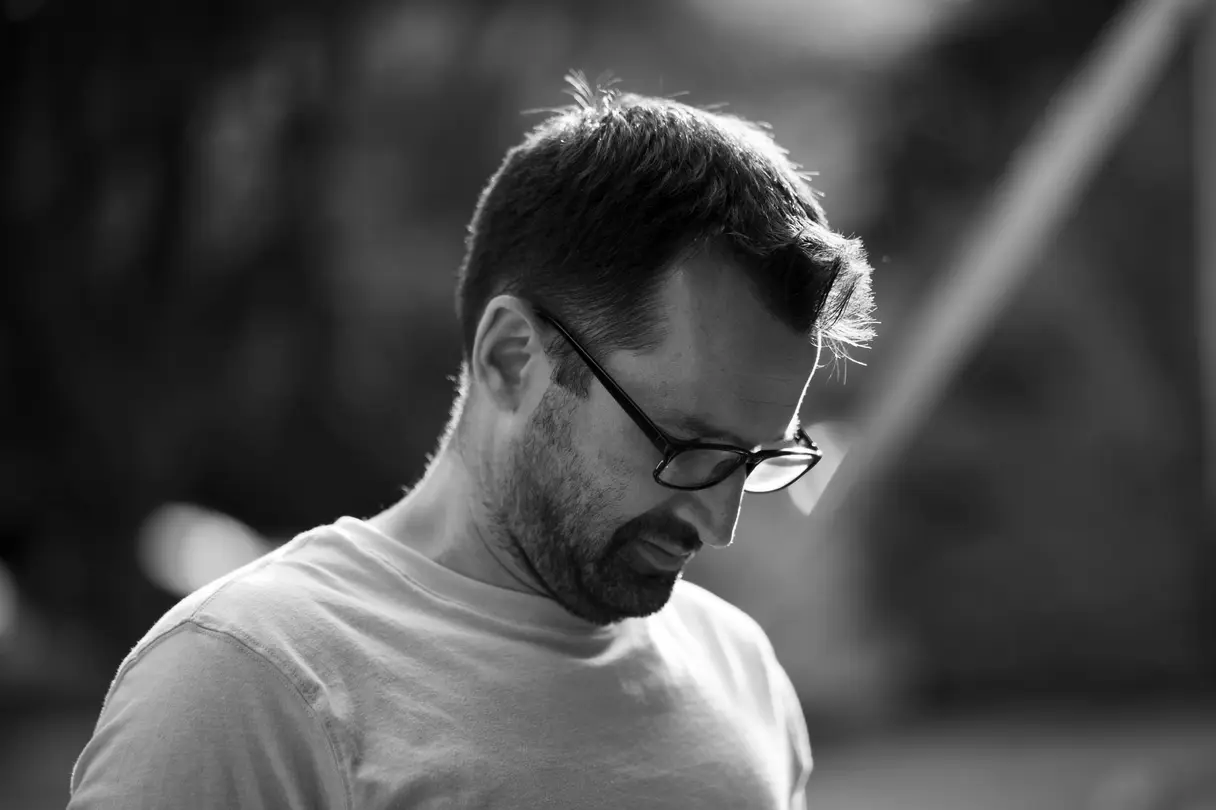 Man in glasses looking down in reflection, backlit by natural sunlight, in black and white for stylistic effect.